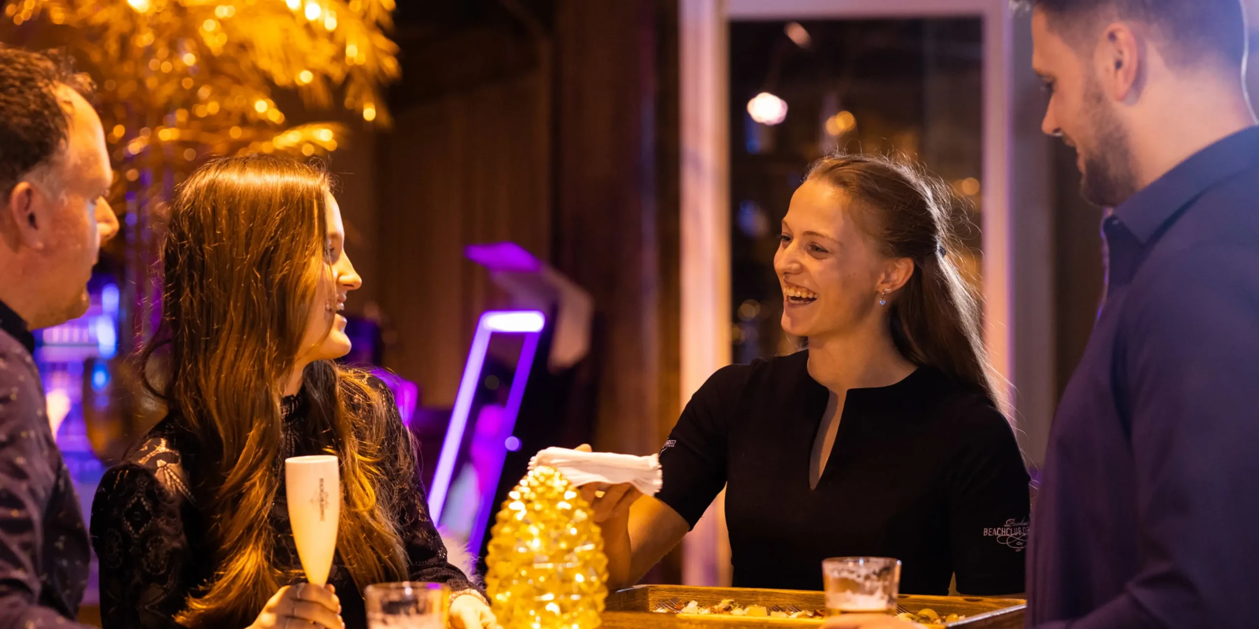 Raumbeduftung-gastgeberin-serviert-getraenke | zaluti b. V. Gastgeberin von beachclub degreez serviert lächelnden gästen getränke bei einer festlichen veranstaltung, bei der raumbeduftung das erlebnis verstärkt.