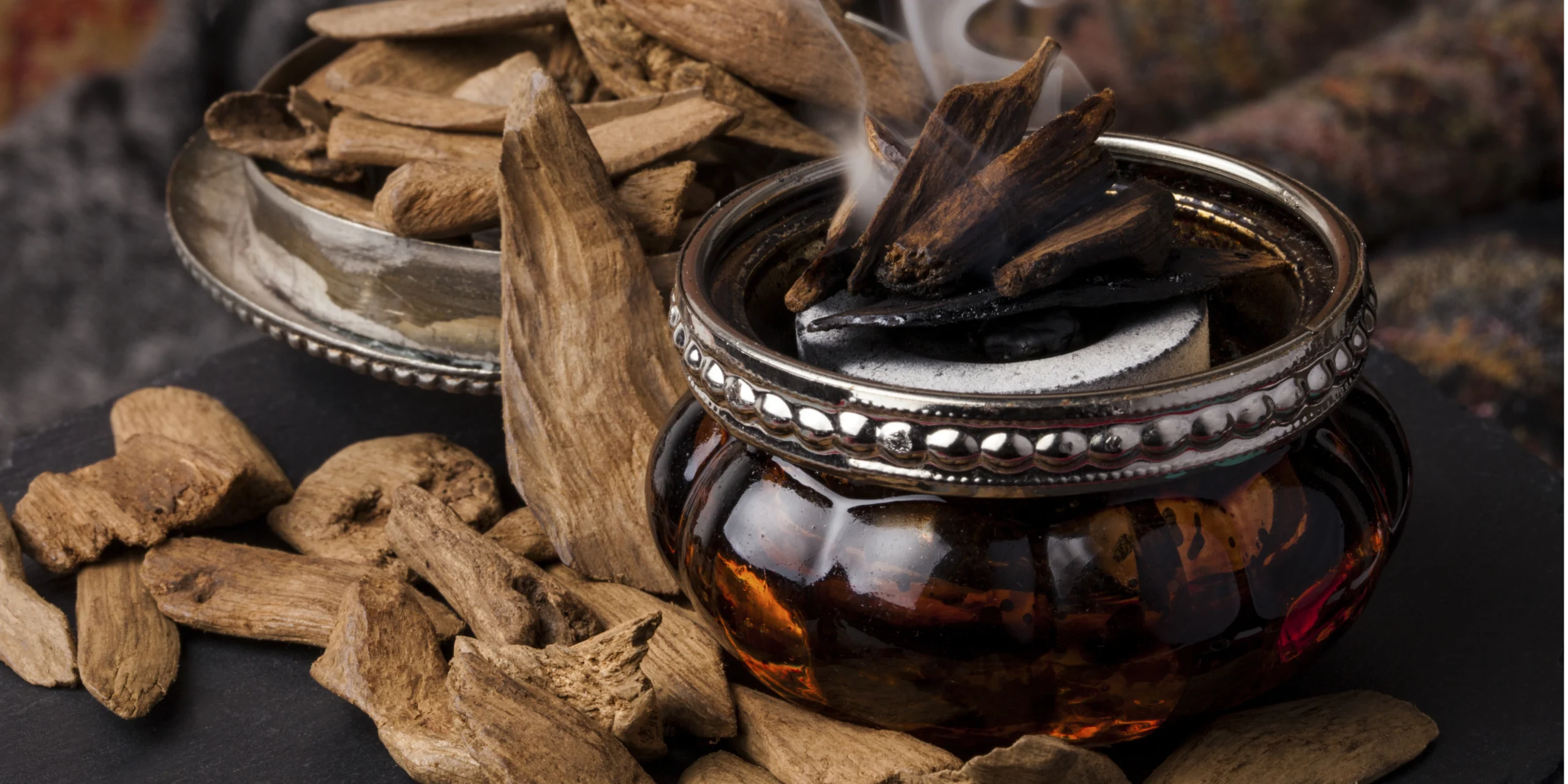 Kleines bernsteinfarbenes räuchergefäß mit brennendem agarholz (oud), aus dem aromatischer rauch aufsteigt, umgeben von losen holzstücken.