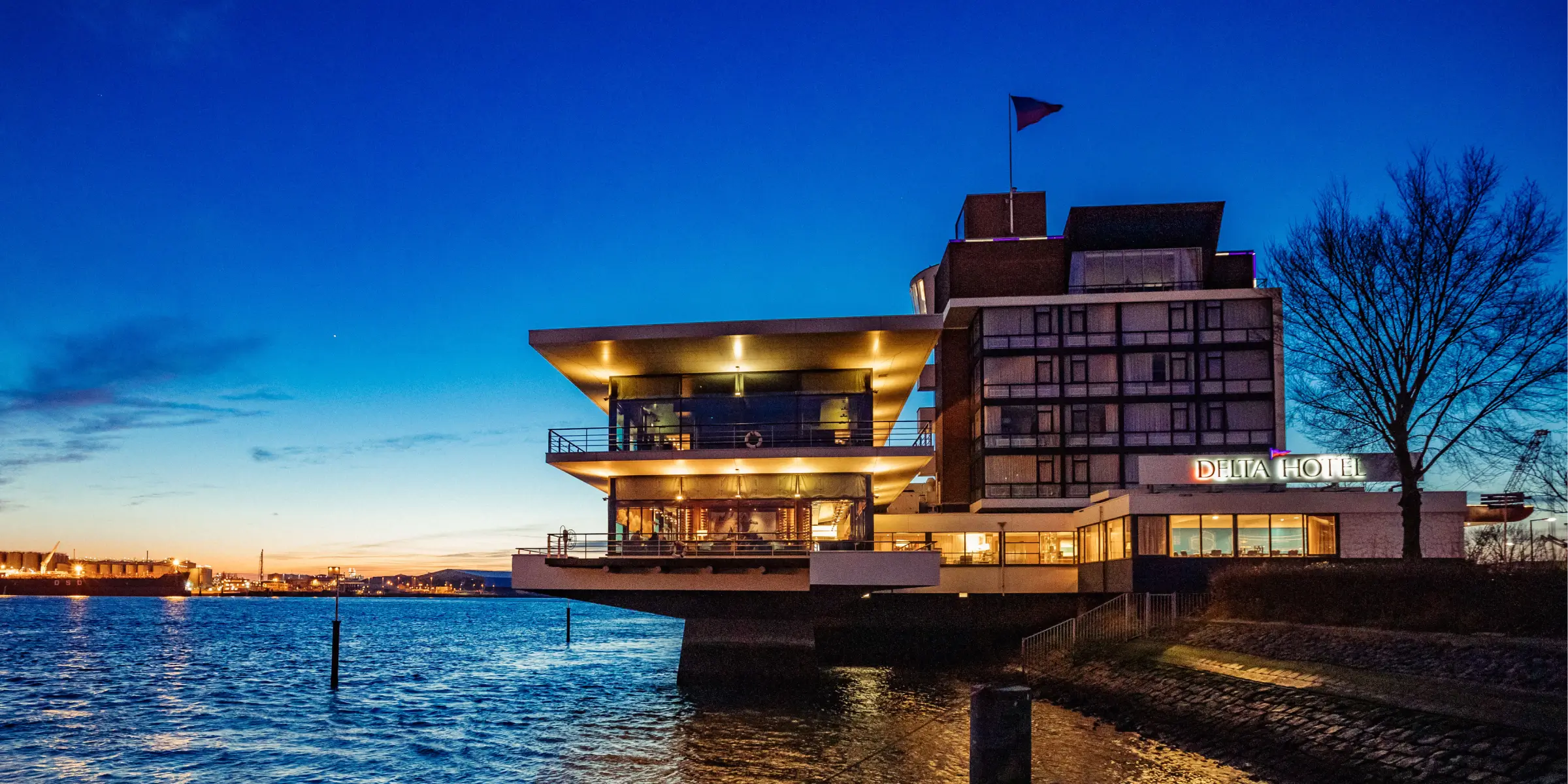 Einzigartige gastfreundschaft am wasser im delta hotel vlaardingen | zaluti b. V. Das delta hotel vlaardingen bei abenddämmerung mit beleuchteter fassade direkt am wasser und blick auf den fluss.
