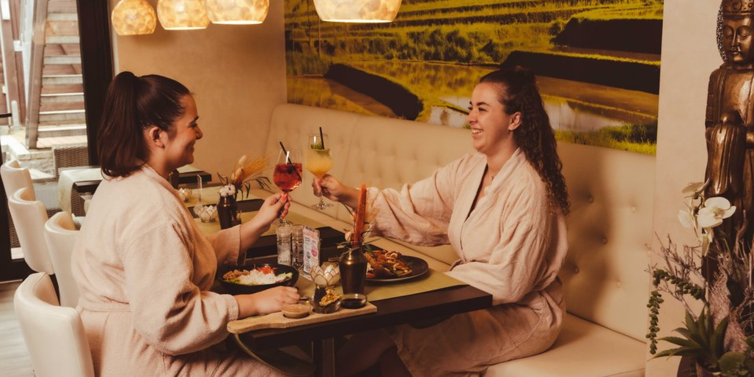 Two women in bathrobes enjoying drinks and a meal in a serene spa lounge setting with soft lighting, a buddha statue, and a calming interior.