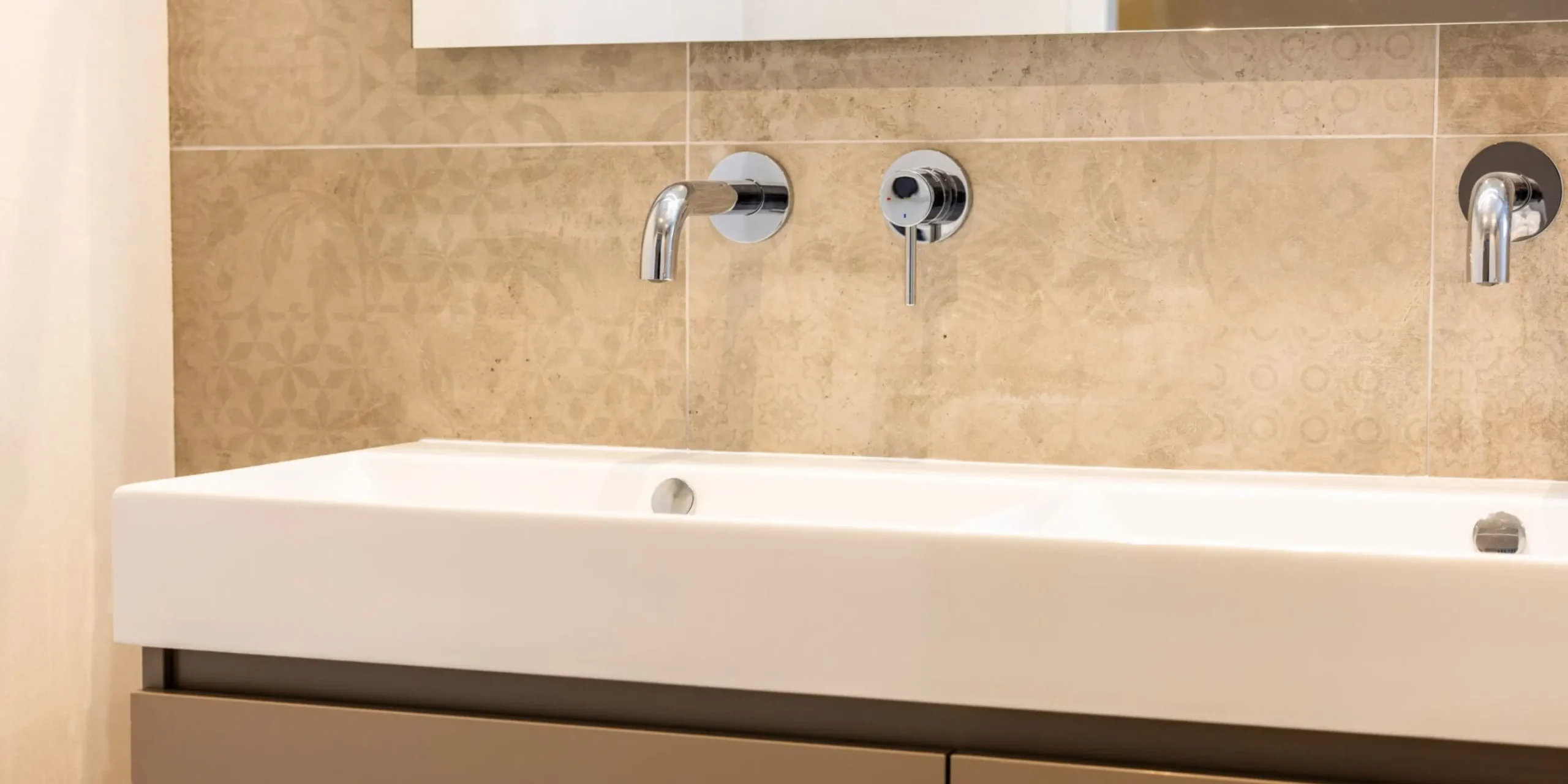 Scent-marketing-showroom-double-sink-detail | zaluti b. V. Close‑up of a modern beige bathroom with a double sink, integrated chrome faucets and a subtle pattern in the wall tiles.