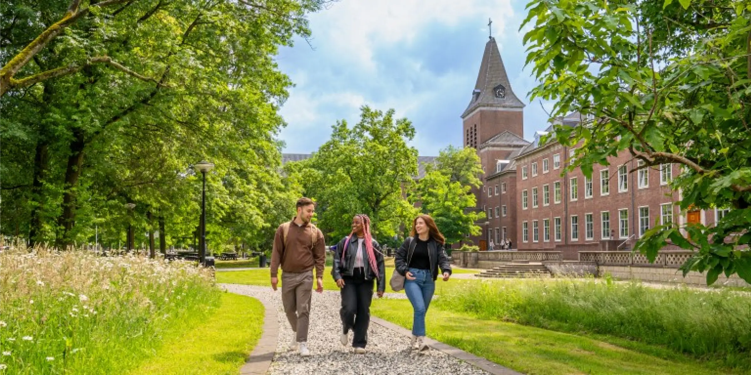 Students walking along a path on the green campus grounds of Breda University of Applied Sciences in Breda, with trees and a historic building in the background.